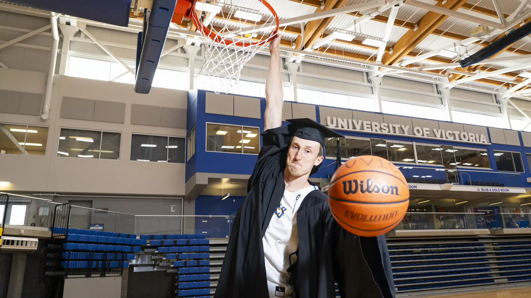 A grad slam dunking a basketball in regalia over his Vikes uniform.