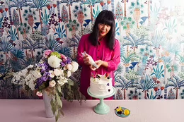 Woman decorating a cake in front of flowery wallpaper.
