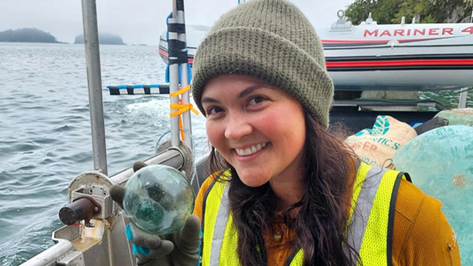 Picture of UVic alum Hannah Gentes smiling to the camera, while holding a glass sphere removed from the ocean. he is on board of a boat during a restoration project excursion.