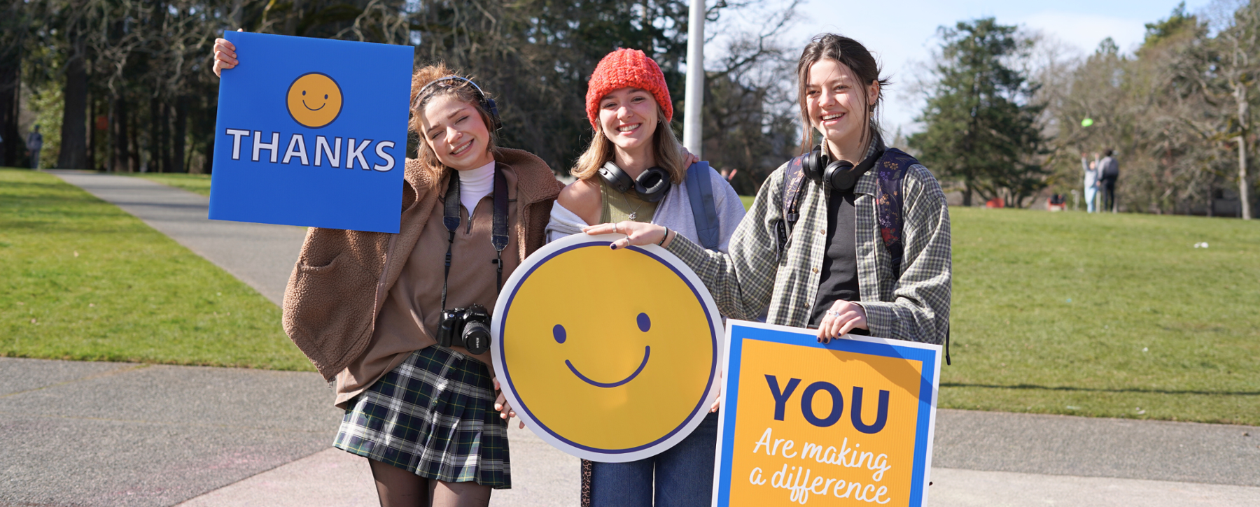 three female students stand together smiling and holding signs, from left to right the signs are "Thanks", a giant yellow smiley, "you are making a difference" 