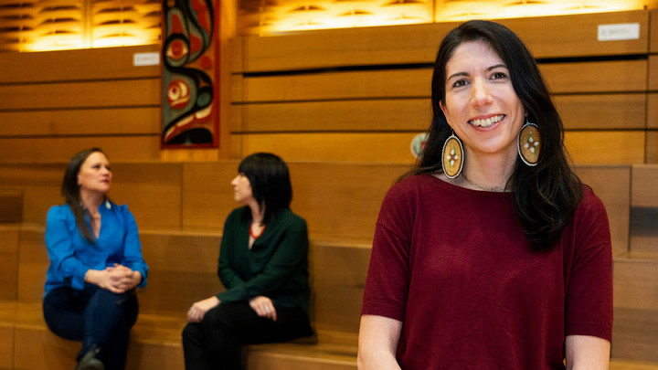 International Women's Day expert Gina Starblanket smiles at the camera with two women sitting and talking in the background.