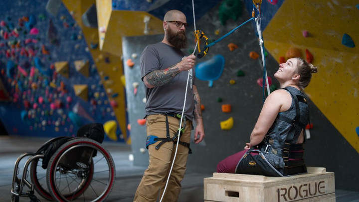 One person standing holding climbing ropes and one person in a sit pulley in front of a climbing wall. There is an empty wheelchair close by them.