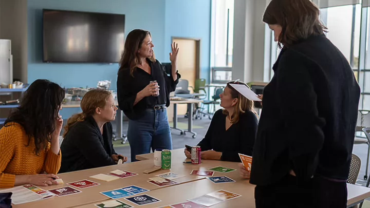 A group of staff sit and stand around a table in discussion. Colourful square cards representing the UN SDGs sit on the table.