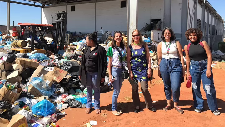 Five women stand together with Jutta Gutberlet standing in the middle. Large piles of trash and a white building are behind them.