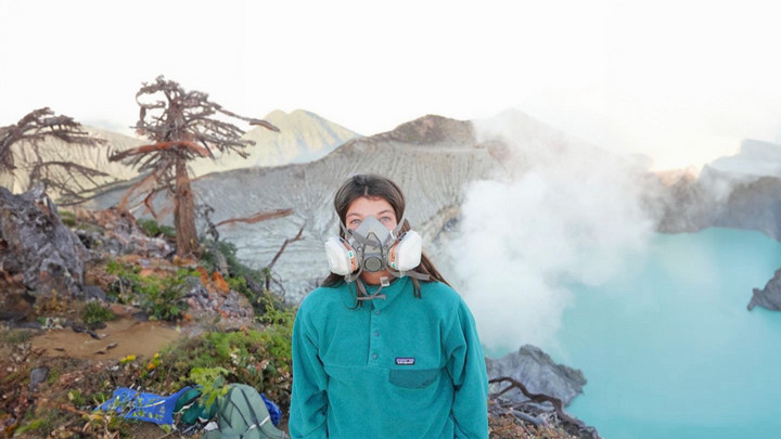 A person wearing a green jacket and a respirator mask stands near a steam-emitting crater lake with a backdrop of rocky terrain and sparse vegetation.