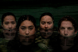 The heads of four young people are half-submerged in water, as the water surface comes up to just below their noses. The individuals are somber and the scene is rainy and green.