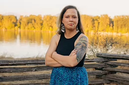 A young woman smiles and stands with her arms crossed in front of a wooden fence, with water in the distance.