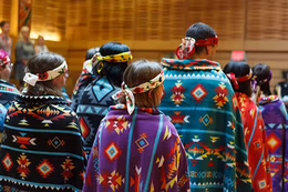 A group of students are shown from the back wearing colourful blankets and head-dresses as part of an Indigenous blanketing ceremony