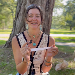 A female student sits on the quad holding a small microphone and piece of paper. There is a tree behind her.