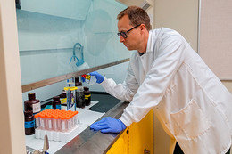 A man wearing blue latex gloves, a white lab coat and glasses leans against a counter in a lab as he pour a liquid into a beaker.