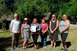 Six people stand together in a line on a sunny day on green grass with trees and a small brown building structure behind them. The woman third from the left holds up a paper document.