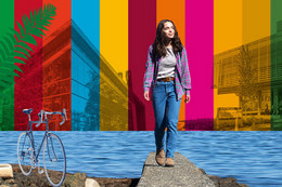 A colour composite with bright coloured stripes, a body of water, a young woman, rocks, tree and a bicycle