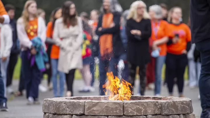 A Sacred Fire burns in a grey brick fire pit as an out of focus crowd of people wearing orange t-shirts stand behind the fire. 