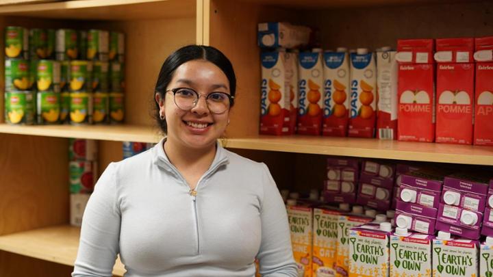 Caro Martinez stands smiling at the camera in front of food bank shelves