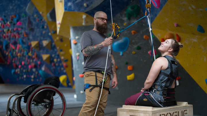 One person standing holding climbing ropes and one person in a sit pulley in front of a climbing wall. There is an empty wheelchair close by them.