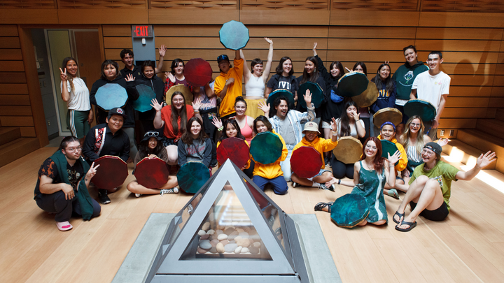 A large group of mini-U students and chaperones smile for the camera in the Ceremonial Hall; many are holding dyed hand drums and there's a grey indoor triangle fire pit in front of them. 