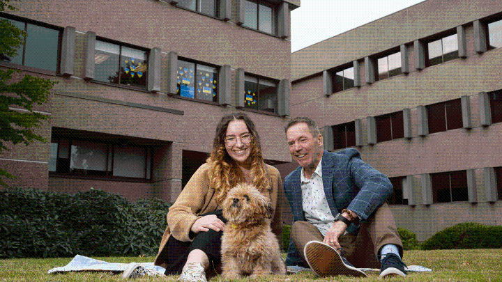 A man and young woman and small dog sitting on grass outside UVic's Clearihue building