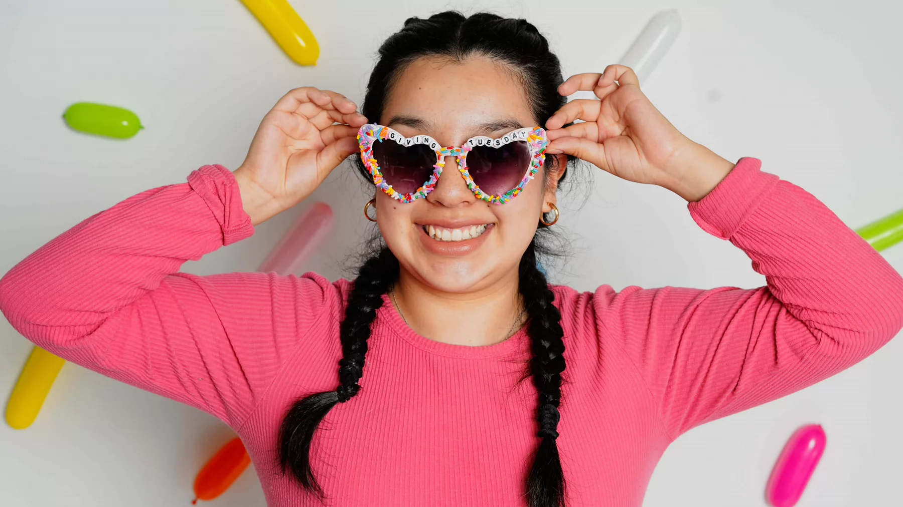 A student wearing heart shaped sunglasses and a bright pink shirt stands in front of a white background with large colourful sprinkles.