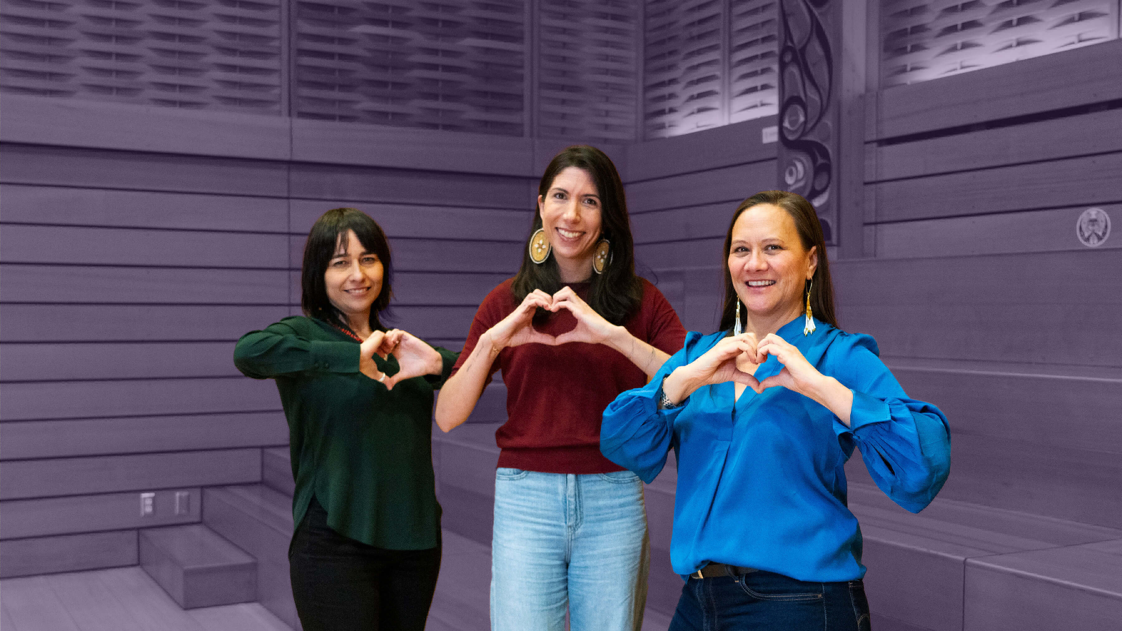 Three women face the camera smiling while giving a heart hand symbol.