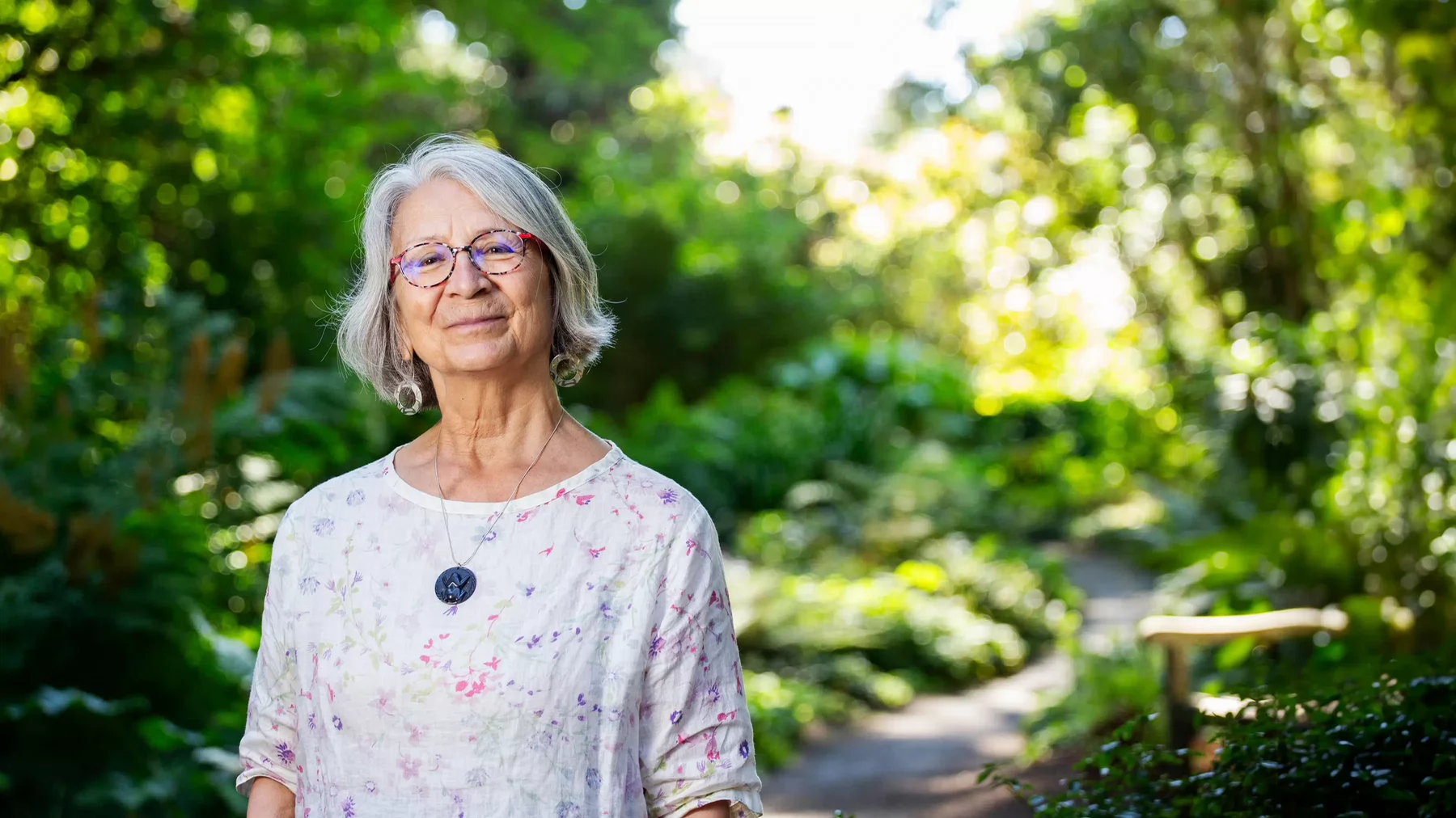 Val Napoleon wears a light white and floral shirt and stands on campus with foliage in the background.