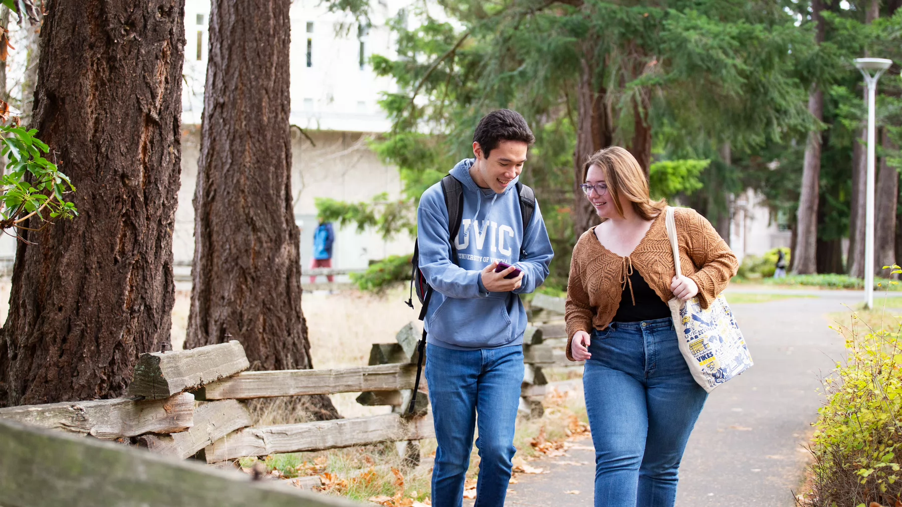 Two UVic students walk down a pathway on campus on a sunny day with the UVic quad in the background. 