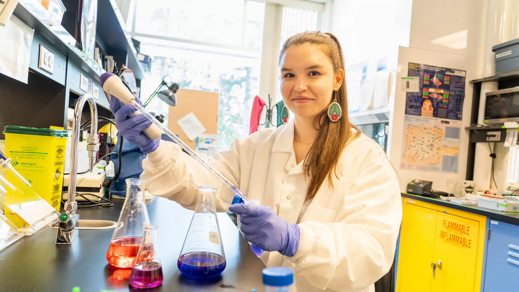 Student wears a white lab coat and blue gloves sits in a lab with colourful liquids in glass containers of different sizes.