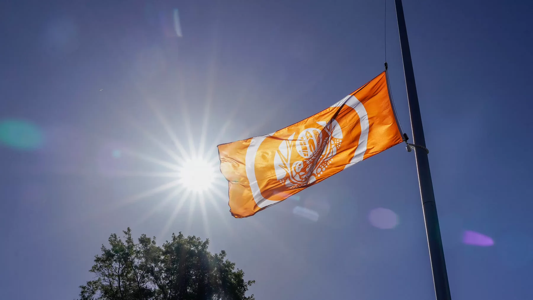 The National Centre for Truth and Reconciliation Survivors’ Flag is seen flying under a sunny sky. The flag is predominantly orange with white artwork depicting feathers, children. 