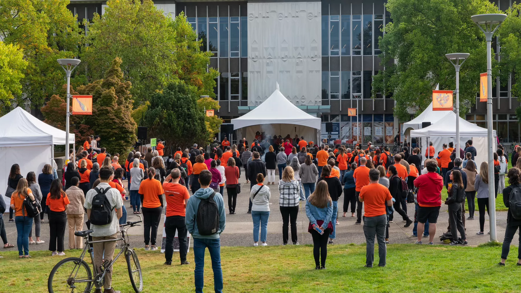 A crowd of people in orange t-shirts are seen from behind as they look towards a stage where smoke from a Sacred Fire can be seen in the air. 