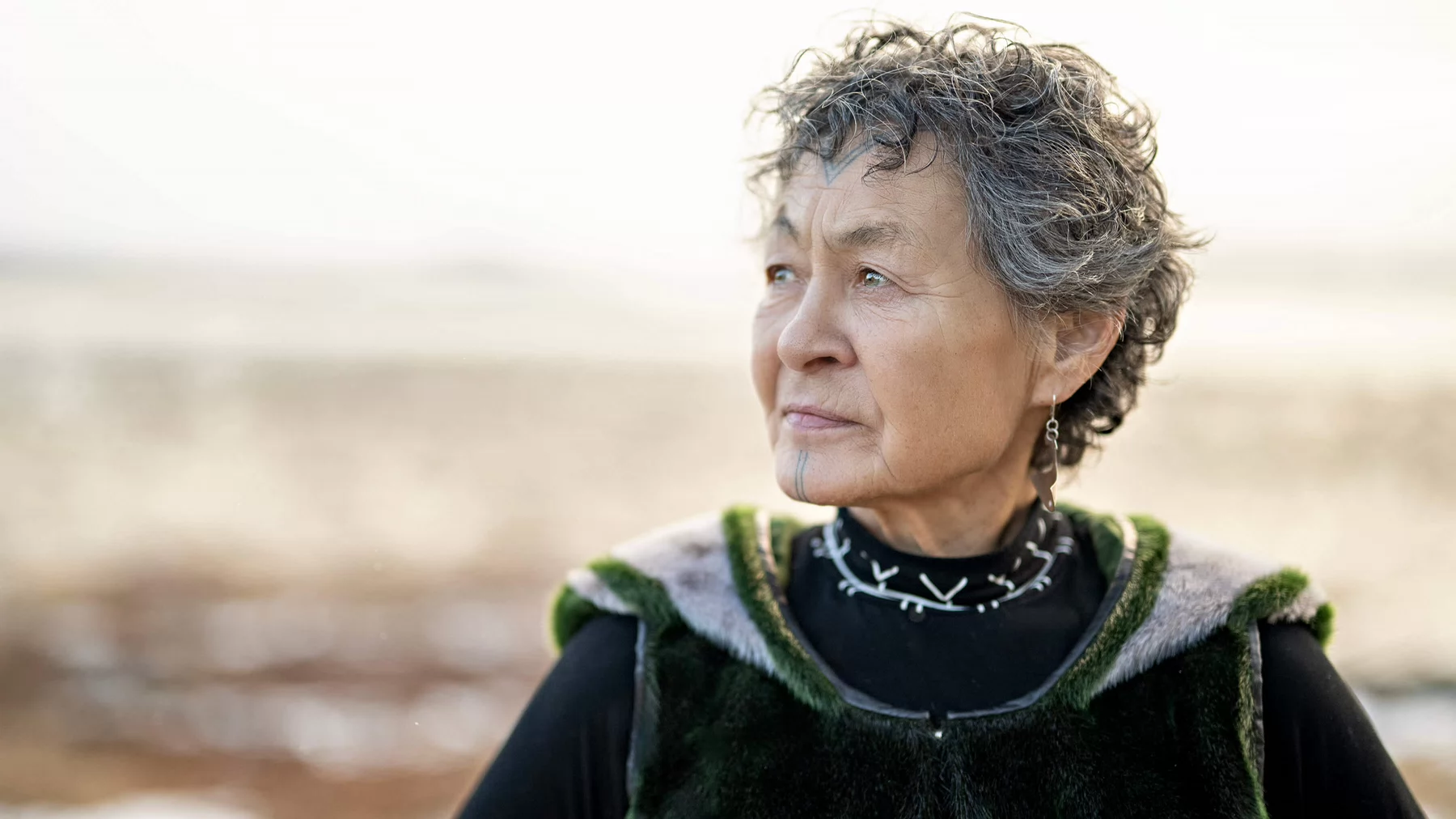 Woman with fur vest and Inuit tattoo marking on her face looking into the distance.