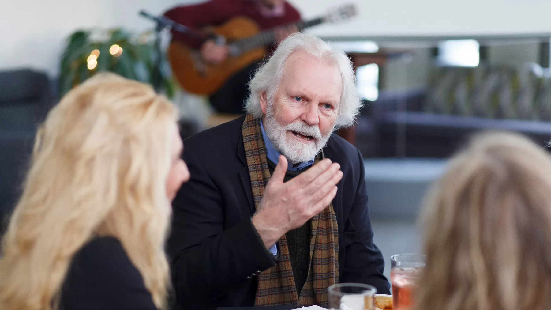 Order of Canada member, Michael Prince sits at a table in discussion with two other people. A person plays the guitar in the background. 