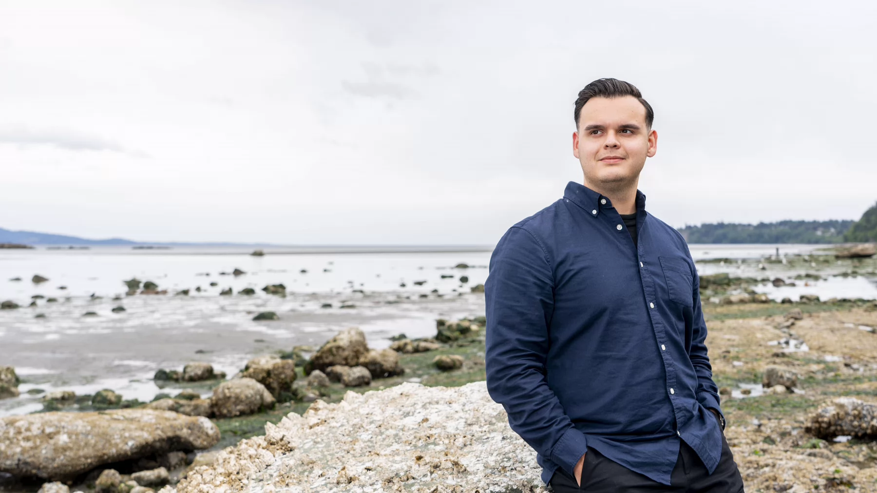 A young Indigenous man wearing a blue shirt stands on the shoreline of Cordova Bay. The tide is out, leaving kelp and barnacle-covered rocks visible in the background.