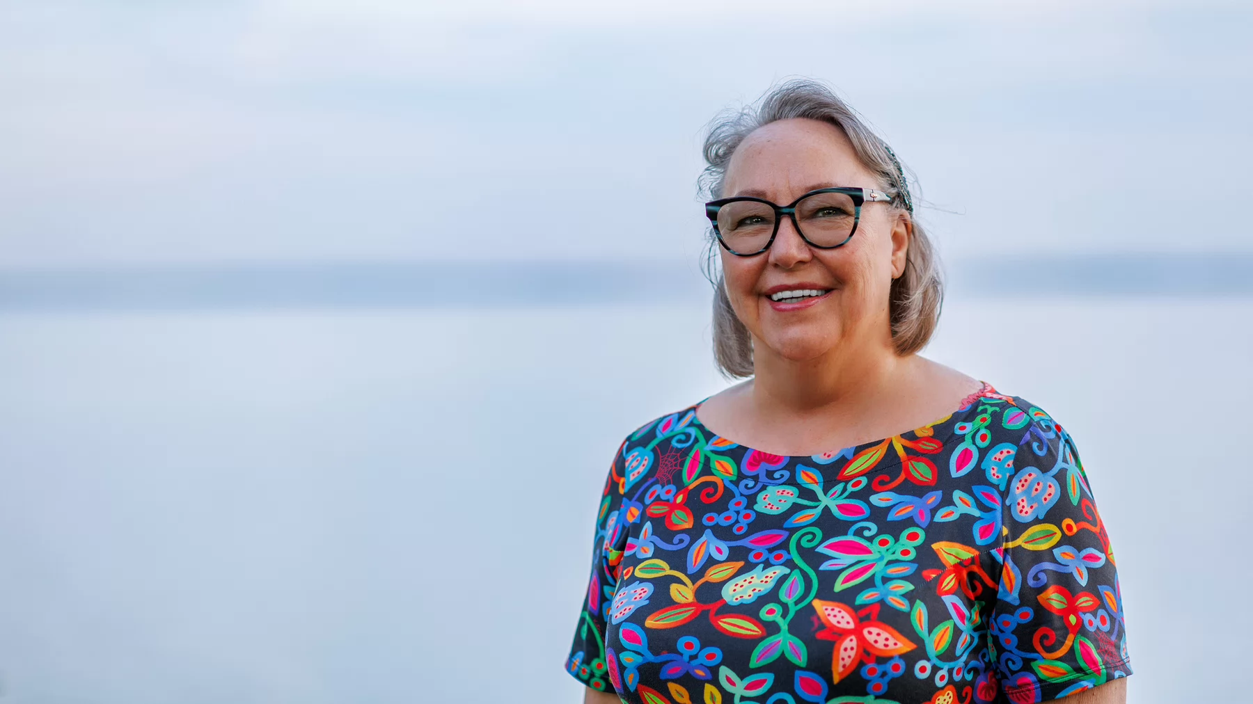 Siila Watt-Cloutier standing in front of a misty ocean wearing glasses and a colourful floral top with Indigenous designs.