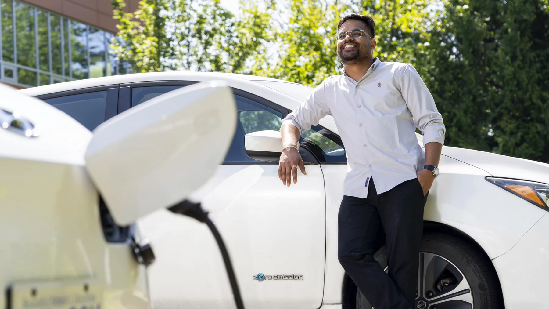UVic student, Akash leans against a white electric vehicle on campus. A second electric vehicle is plugged in, charging, in front of him. 