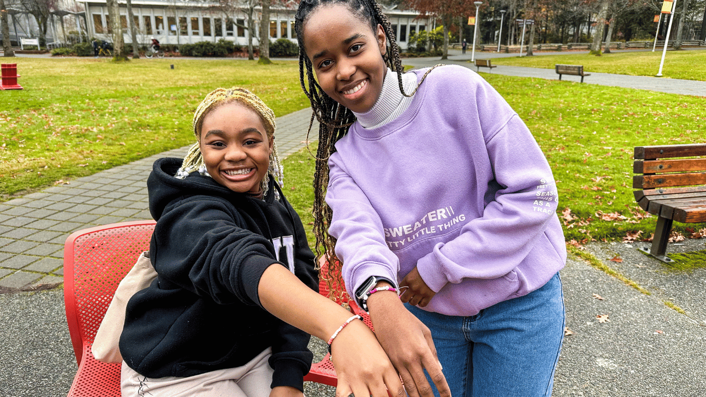 Two students smile at the camera with their arms outstretched showing off colorful beaded bracelets. 