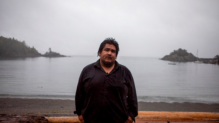 Darrell WIlliams Jr. stands facing the camera on a Yuquot inlet, with boats and land masses fading into the ocean behind him