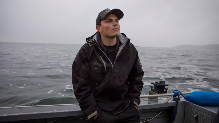 Riley Richardson leans against the hull of a boat with the open ocean behind him