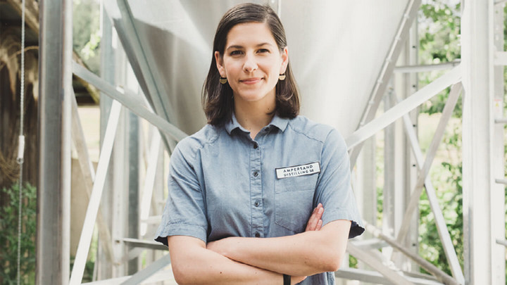 Woman with shoulder length hair standing with arms crossed and wearing a button up work short with a patch that reads "Ampersand Distilling"