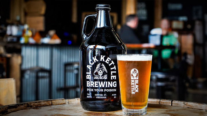 Glass of beer  that says Black Kettle beside a growler that says Black Kettle Brewing Pick Your Poison atop a barrel in a brewery tasting room.