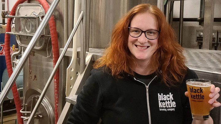 Woman with shoulder length hair wearing a Black Kettle Brewing sweat shirt holding a glass of beer with beer kegs behind her.