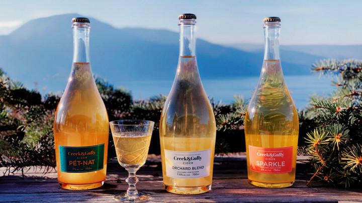 Three clear bottles of cider that say Creek & Gully overlooking a lake.