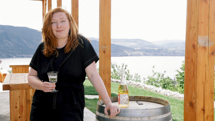 Woman holding a glass of cider poured from a bottle in front of a lake. 