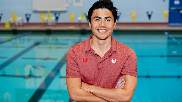 Man in red polo shirt standing arms crossed and smiling in front of an indoor swimming pool.