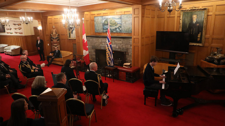 A youth performing on a piano for a small audience wearing military uniforms in a room with a fireplace and flags of Canada and British Columbia..