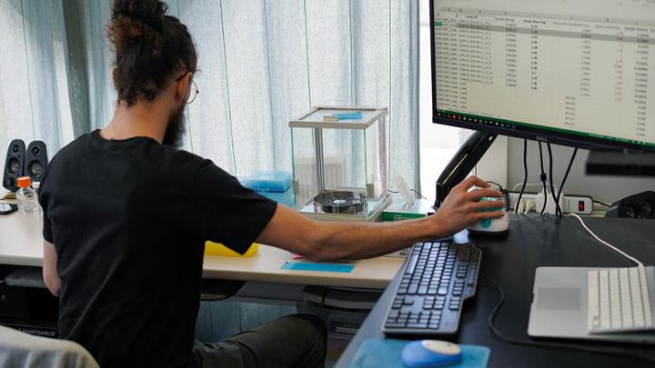 Man in a ponytail places a vial containing an unknown substance into a testing machine hooked up to a computer. 