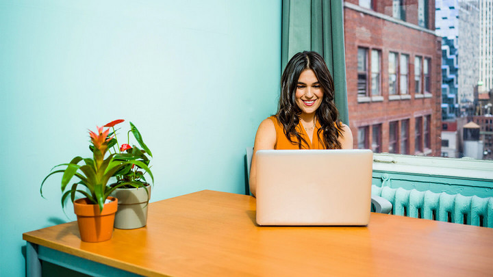 Women smiling and sitting at a table while working on her laptop in front of a window with red brick building outside.
