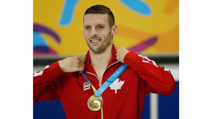 Man in red track suit top with maple leaf logo and medal around his neck smiling. 