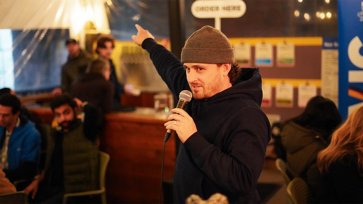 Man wearing a toque speaks into a microphone while pointing. 