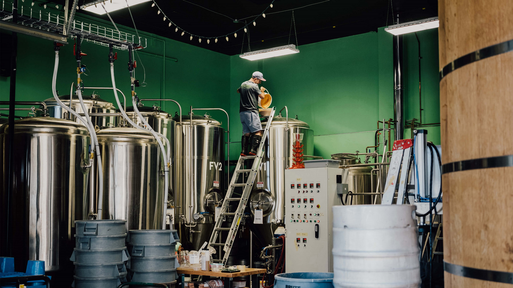 Man in baseball hat, shorts and rubber boots stands on a ladder pouring contents of a bucket into a tall beer tank.