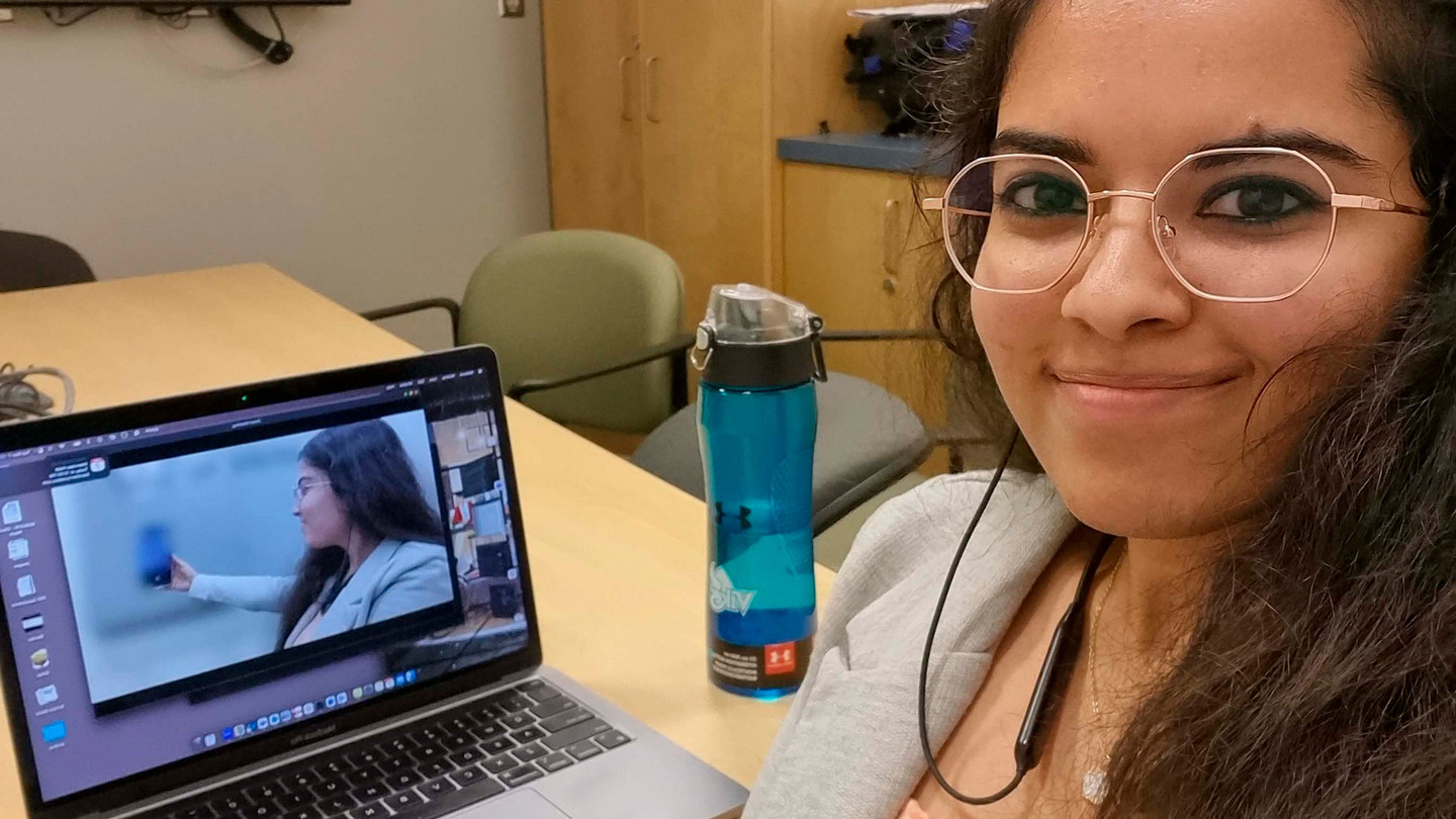 A student with glasses and long hair sits at a desk in front ot a laptop that has a video of the same student showing off a Blackberry product.