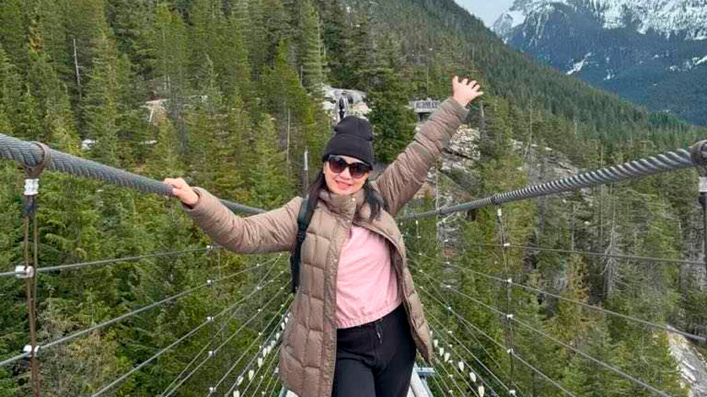 A student wearing a toque and winter jacket stands on a rope bridge with mountains behind her.